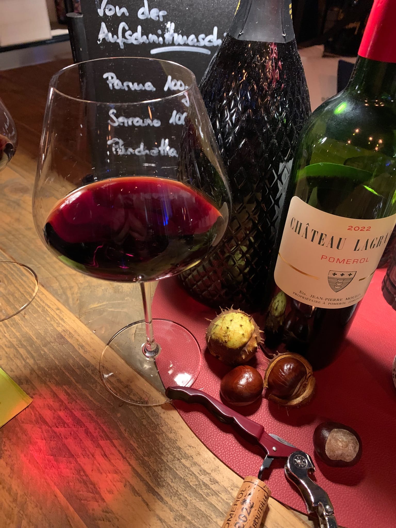 Rotweinglas vor der Aufschnitt-Kreidetafel, daneben Château Lagrange Pomerol 2022 und Kastanien